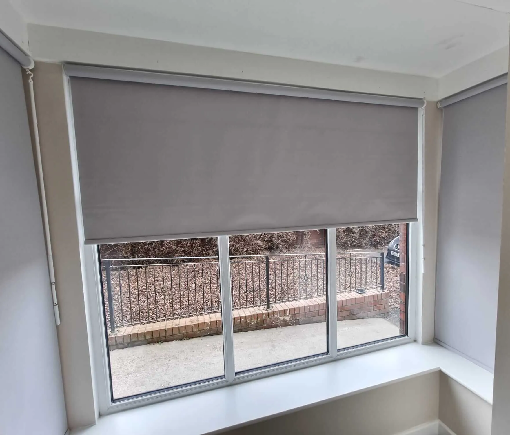 Two dim-out roller blinds fitted to a wide bay window at Beech House Care Home with railings and garden beyond