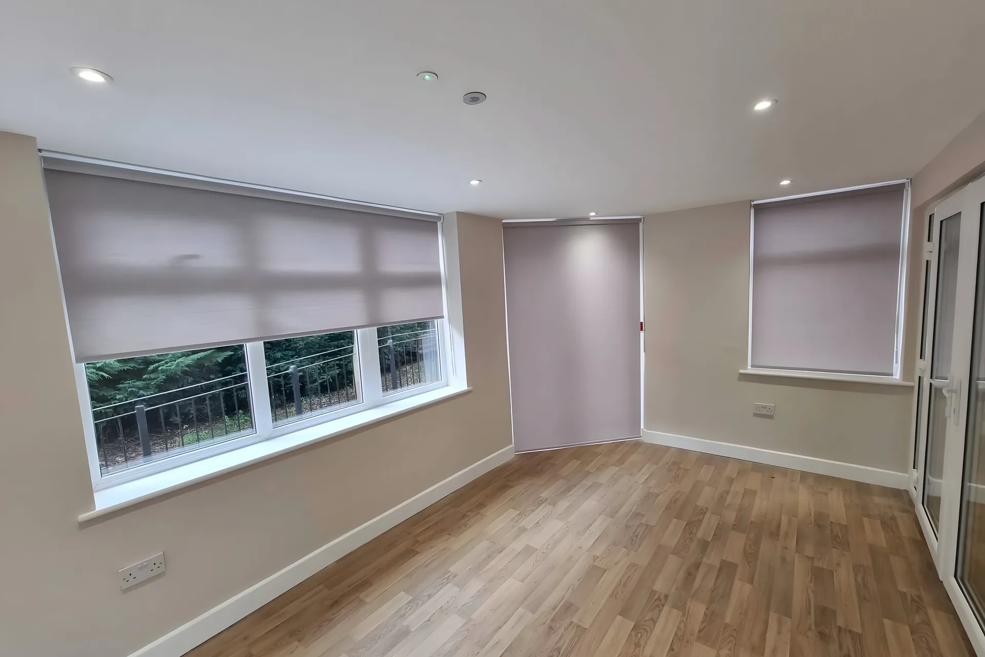 Dim-out roller blinds half lowered across a run of windows in a bright day room at Beech House Care Home, wood-effect floor below