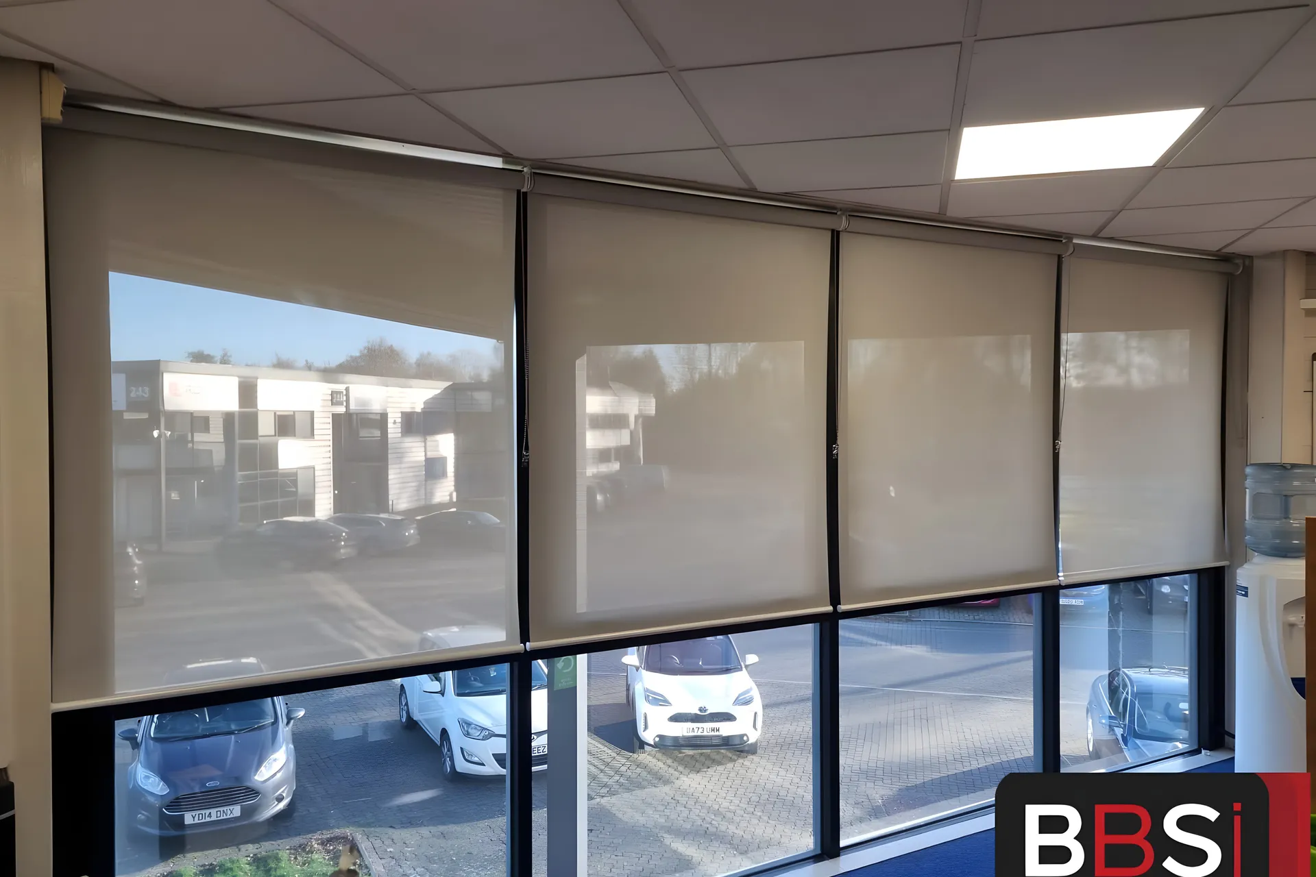 Screen fabric roller blinds lowered across a glazed corner of the Building Technology Systems office, car park and neighbouring units visible through the weave