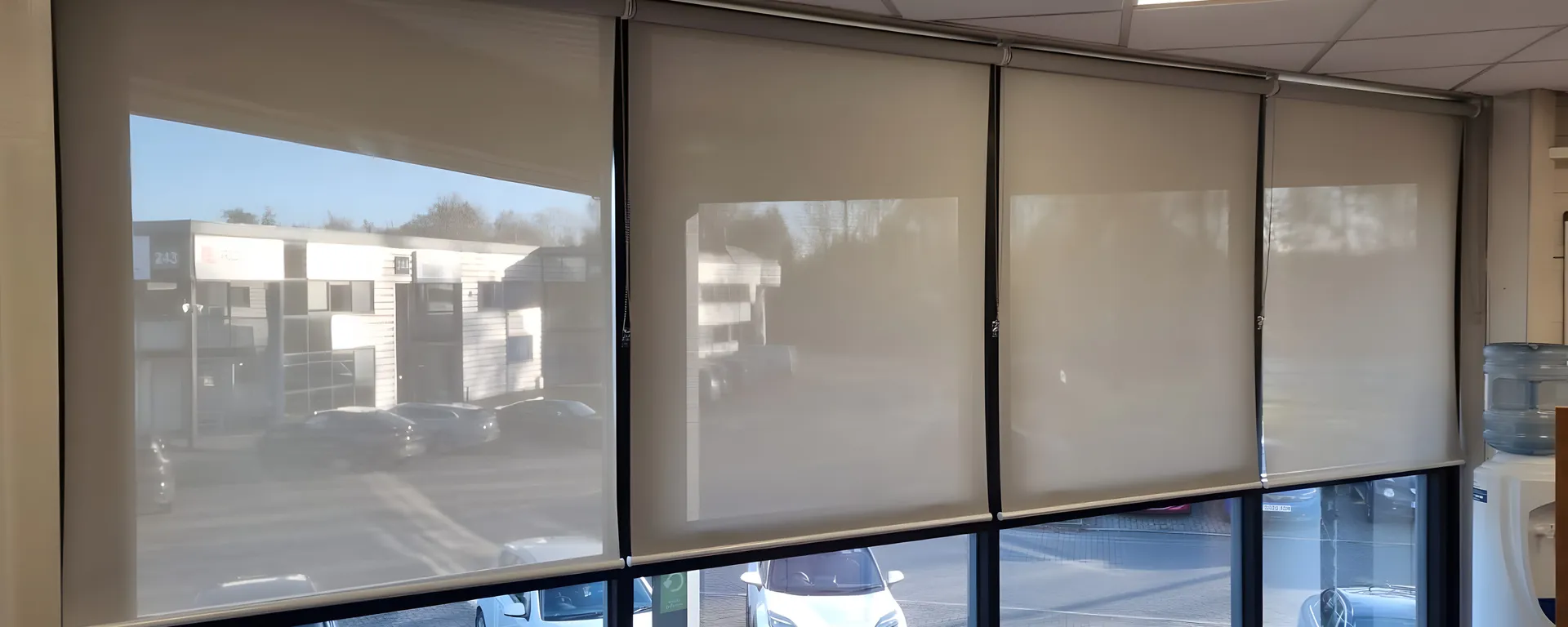 Screen fabric roller blinds lowered across a glazed corner of the Building Technology Systems office, car park and neighbouring units visible through the weave