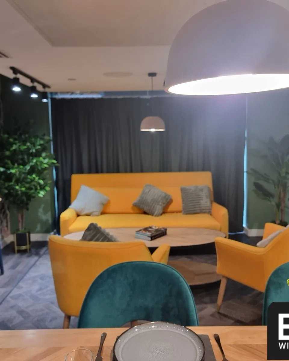 Dark curtains drawn across the full window wall of a shared lounge at Cale Cross, with yellow sofas and a dining table in front