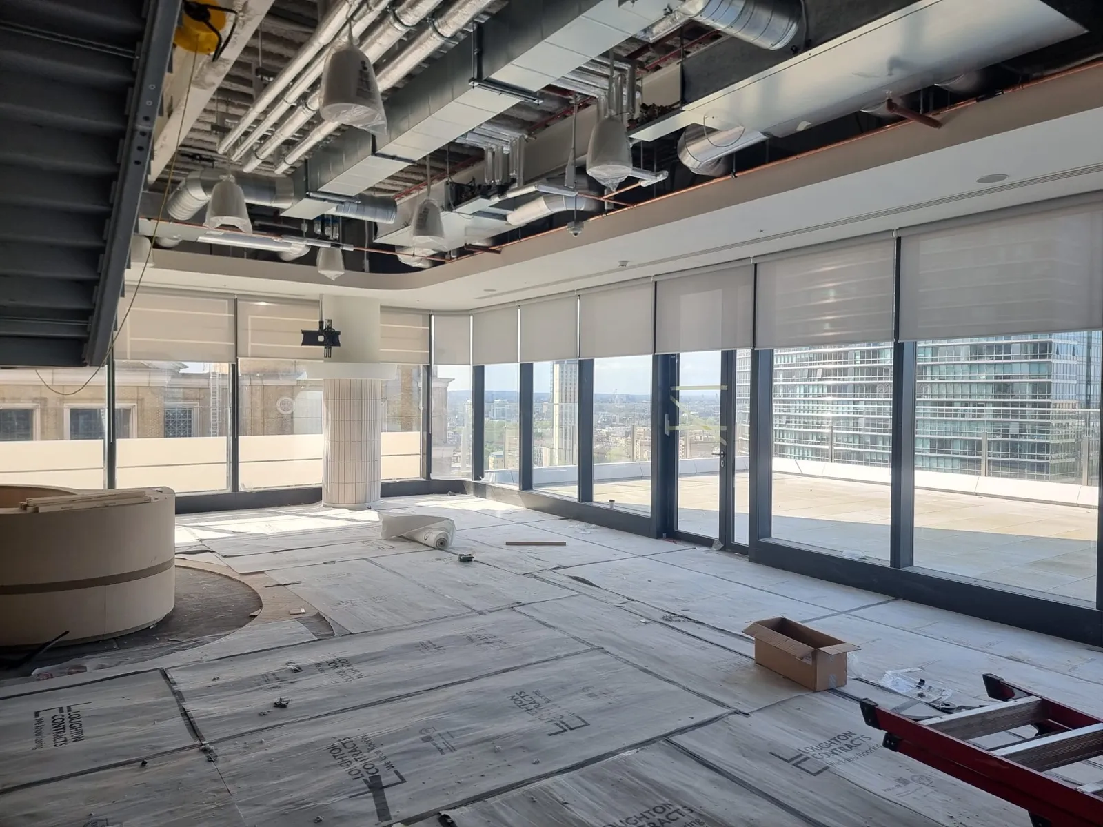 Screen fabric roller blinds along the curtain wall of a Canary Wharf office floor mid fit-out, floor protection down and joinery still going in