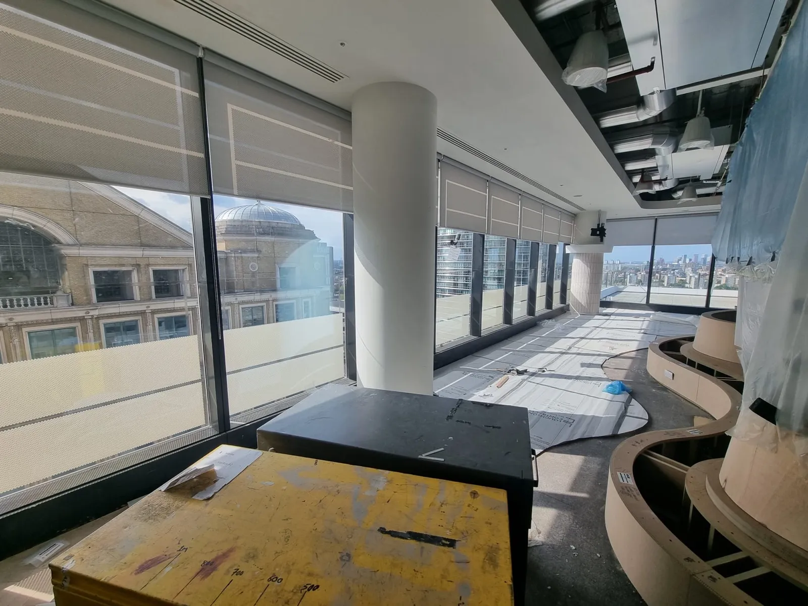 A run of screen fabric roller blinds around the perimeter glazing and terrace doors of an open office floor at Canary Wharf during the fit-out