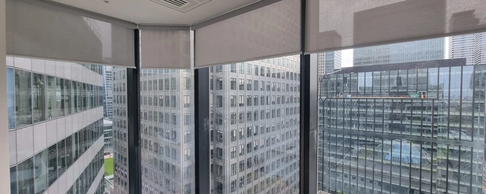 Screen fabric roller blinds on each faceted pane of a curved curtain wall corner at Canary Wharf, raised part way with neighbouring towers and a roof garden below