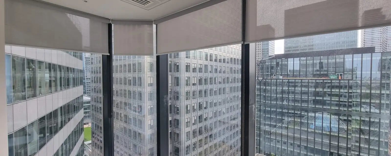 Screen fabric roller blinds on each faceted pane of a curved curtain wall corner at Canary Wharf, raised part way with neighbouring towers and a roof garden below