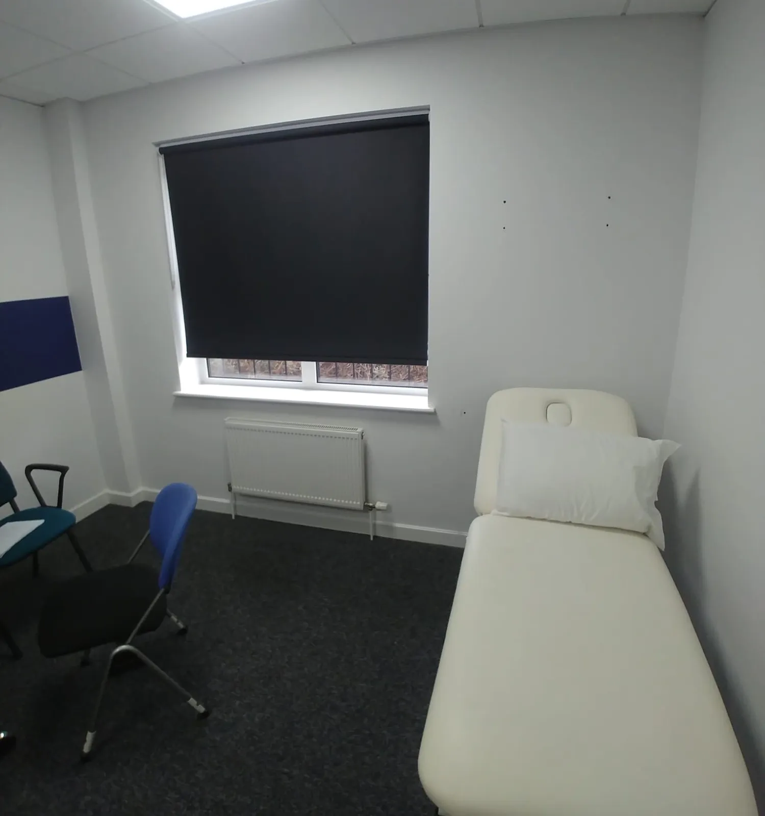 Dark roller blind on an external treatment-room window at a private clinic in Cheadle, with a cream treatment couch and blue chairs