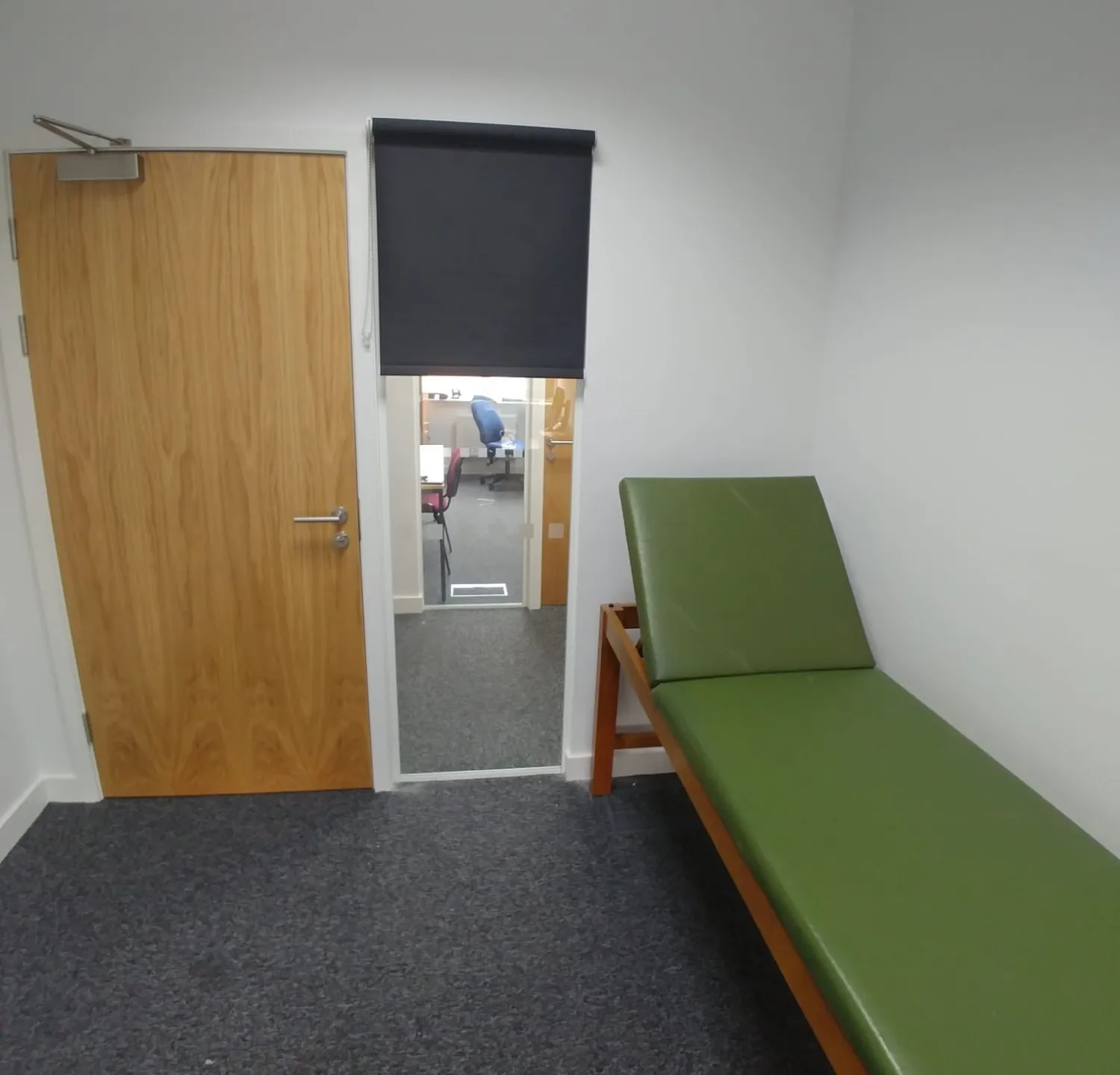 Dark roller blind over the internal glazed panel beside a treatment room door at a private clinic in Cheadle, examination couch in the foreground