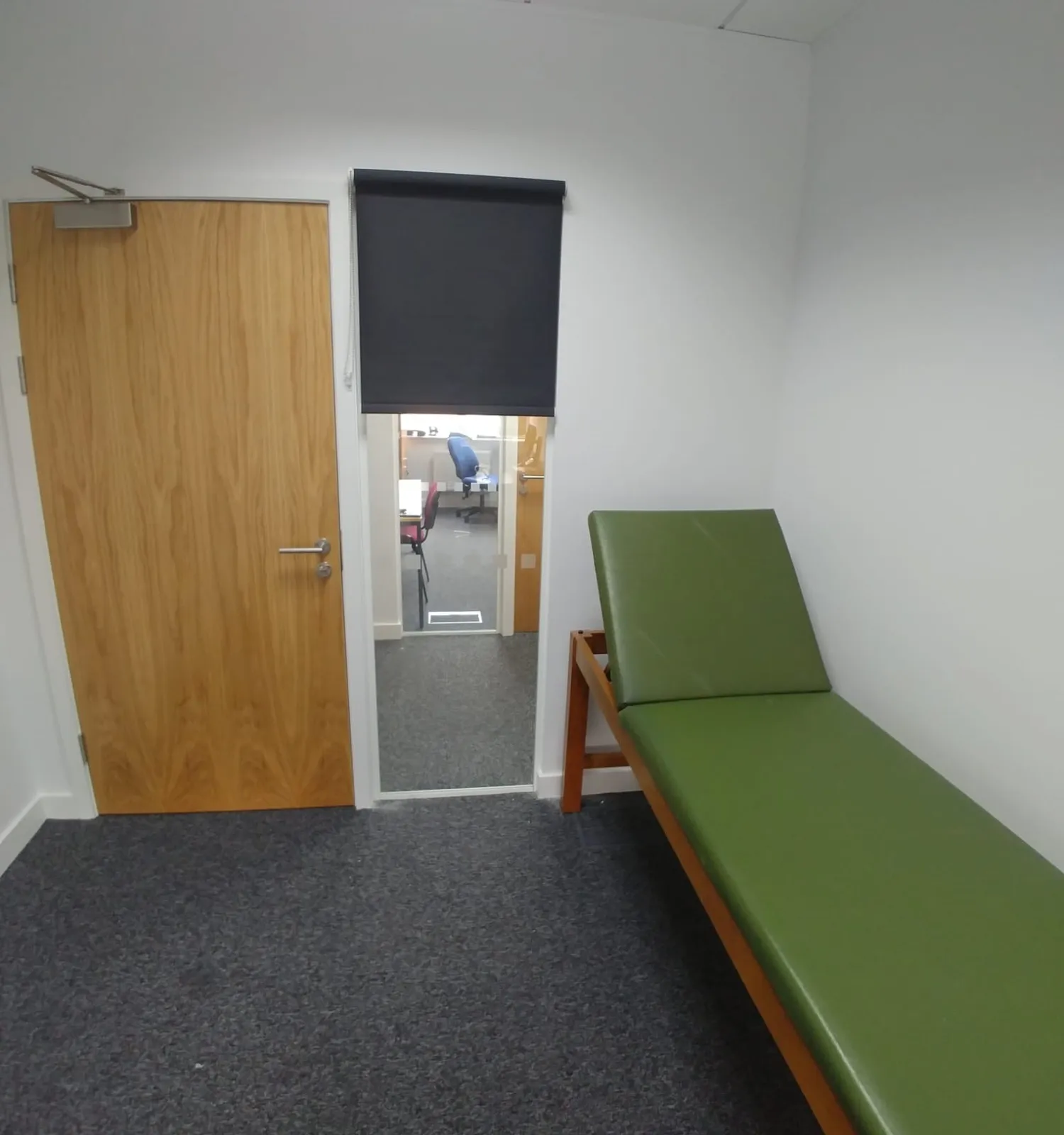 Dark roller blind on an internal glazed panel looking onto the corridor at a private clinic in Cheadle, with a green treatment couch