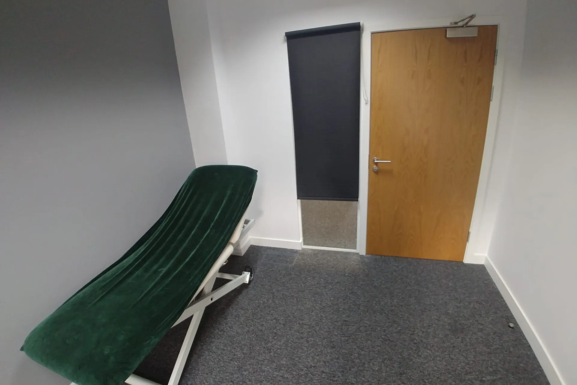 Dark roller blind covering an internal glazed panel next to a treatment-room door at a private clinic in Cheadle, with a green treatment couch in the foreground