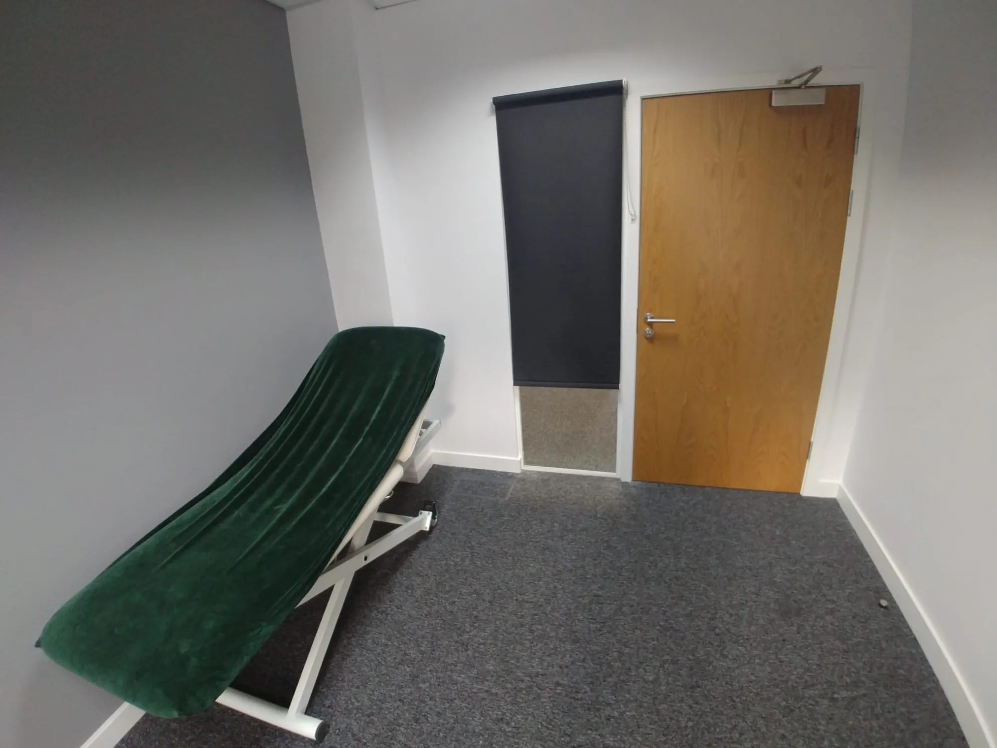 Dark roller blind covering an internal glazed panel next to a treatment-room door at a private clinic in Cheadle, with a green treatment couch in the foreground