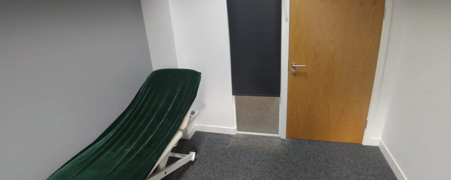 Dark roller blind covering an internal glazed panel next to a treatment-room door at a private clinic in Cheadle, with a green treatment couch in the foreground
