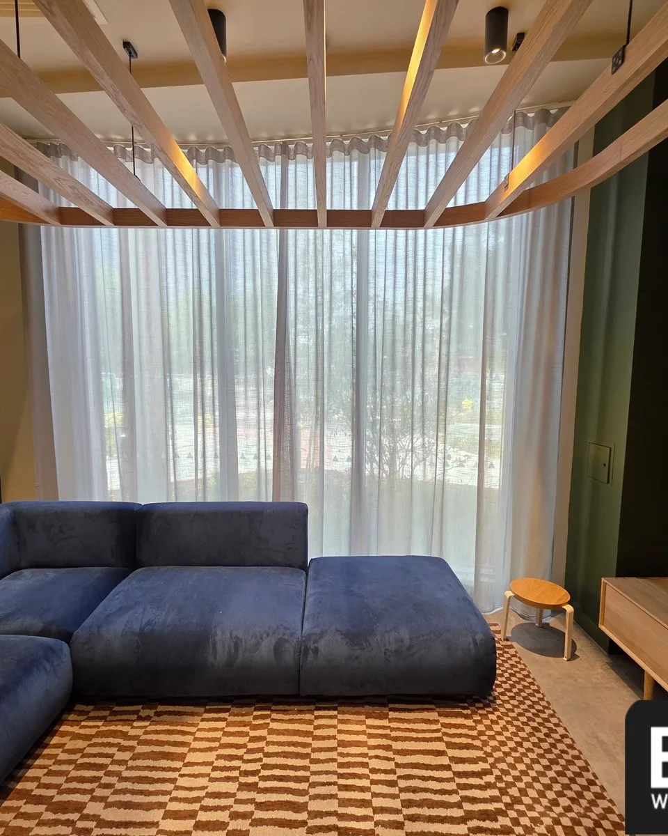 Full height sheer wave curtains across a lounge window behind a blue corner sofa and timber ceiling slats