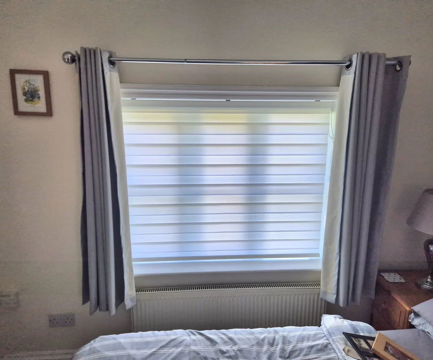 White day and night blind in a bedroom, with grey curtains drawn to either side of the window