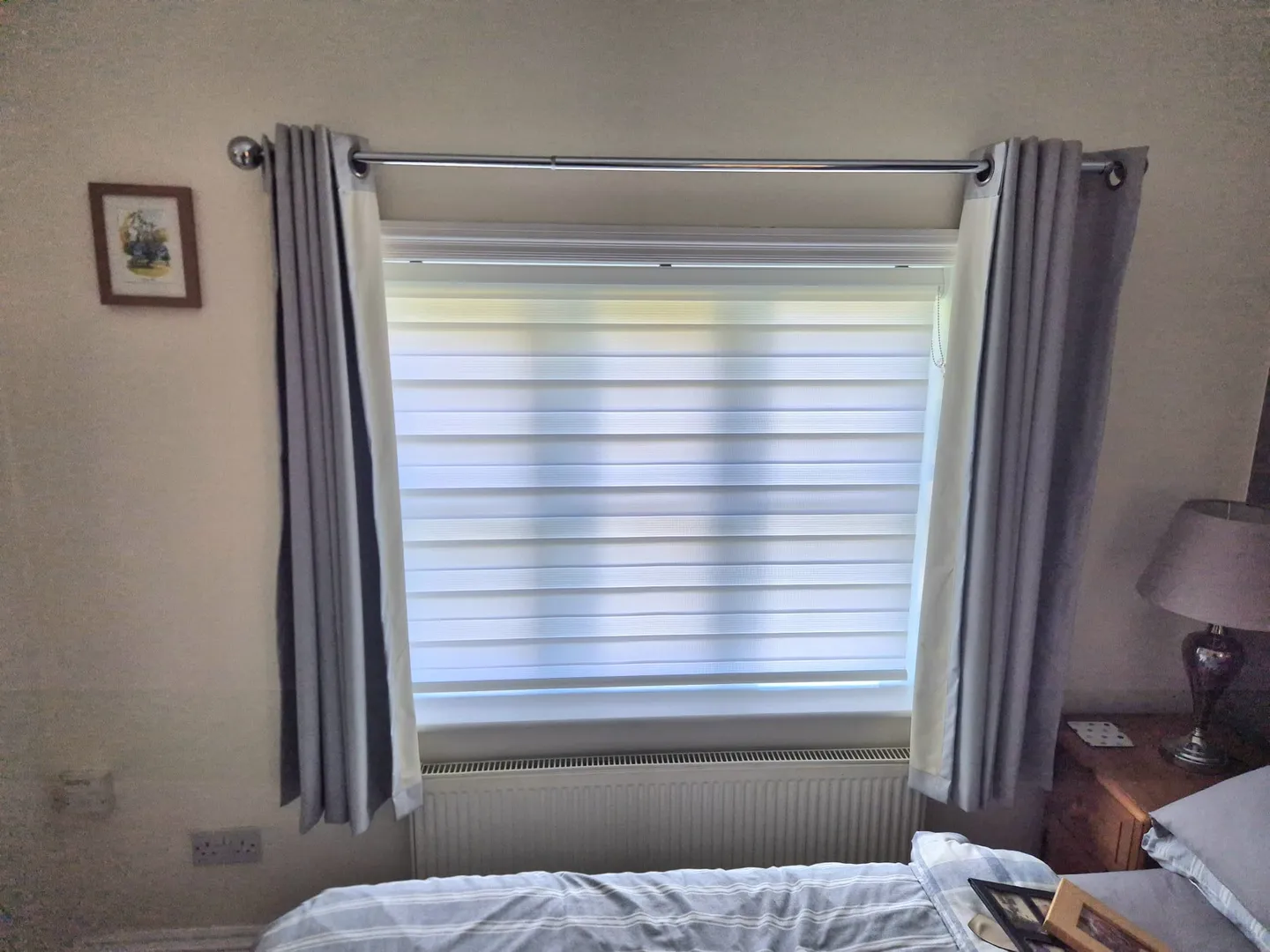 White day and night blind in a bedroom, with grey curtains drawn to either side of the window