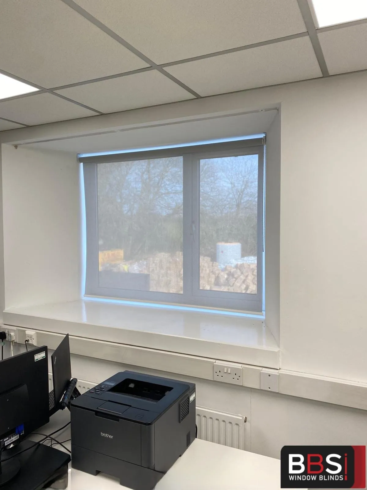 Pale screen fabric roller blind in a recessed office window at Faith House, Radcliffe, above a desk with a printer
