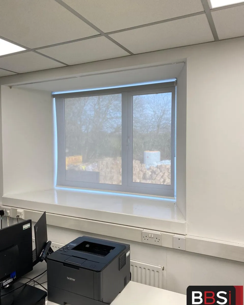 Pale screen fabric roller blind lowered in an office window recess at Faith in Nature, Radcliffe, trees still visible through the weave