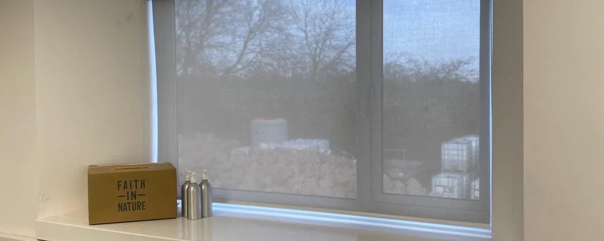 Pale screen fabric roller blind in a recessed office window at Faith House, Radcliffe, with a Faith in Nature box and two silver dispensers on the sill