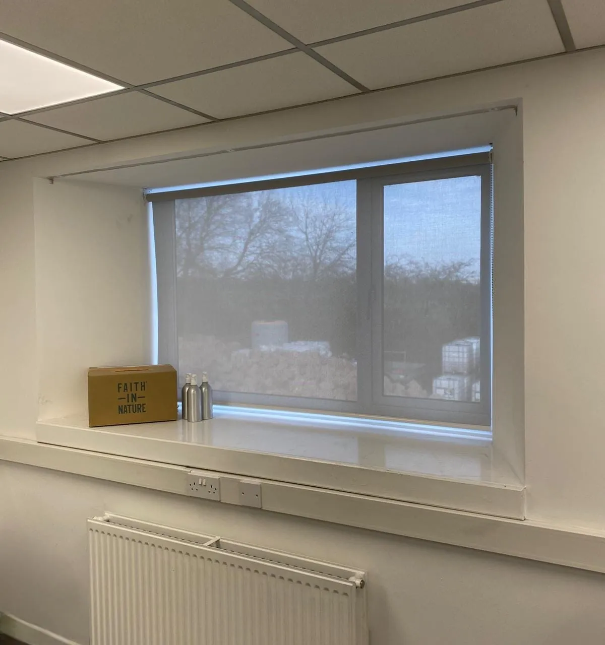 Pale screen fabric roller blind in a recessed office window at Faith House, Radcliffe, with a Faith in Nature box and two silver dispensers on the sill
