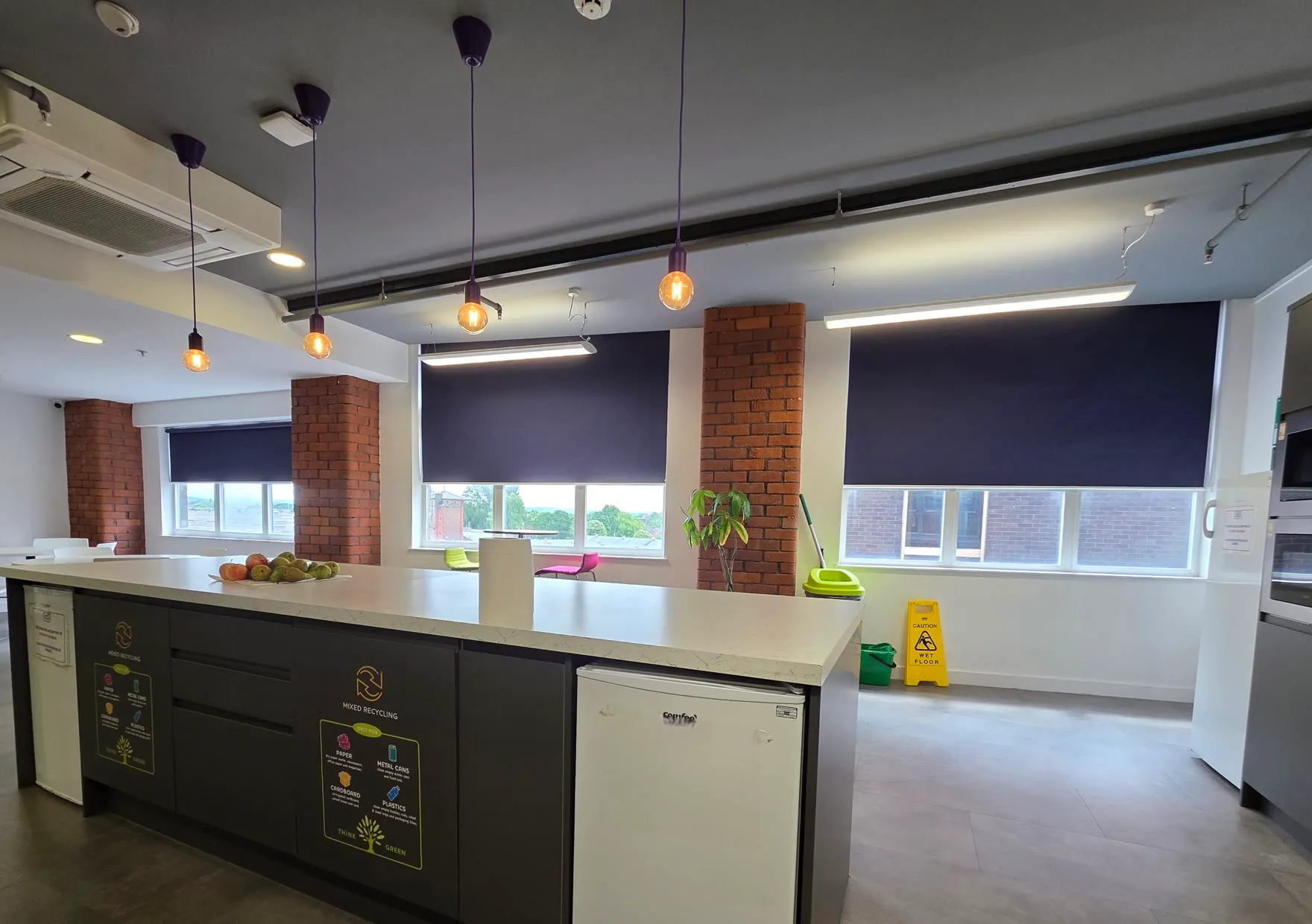 Large navy roller blinds on the staff kitchen windows at Global Banking School, between exposed brick columns, with a kitchen island in front