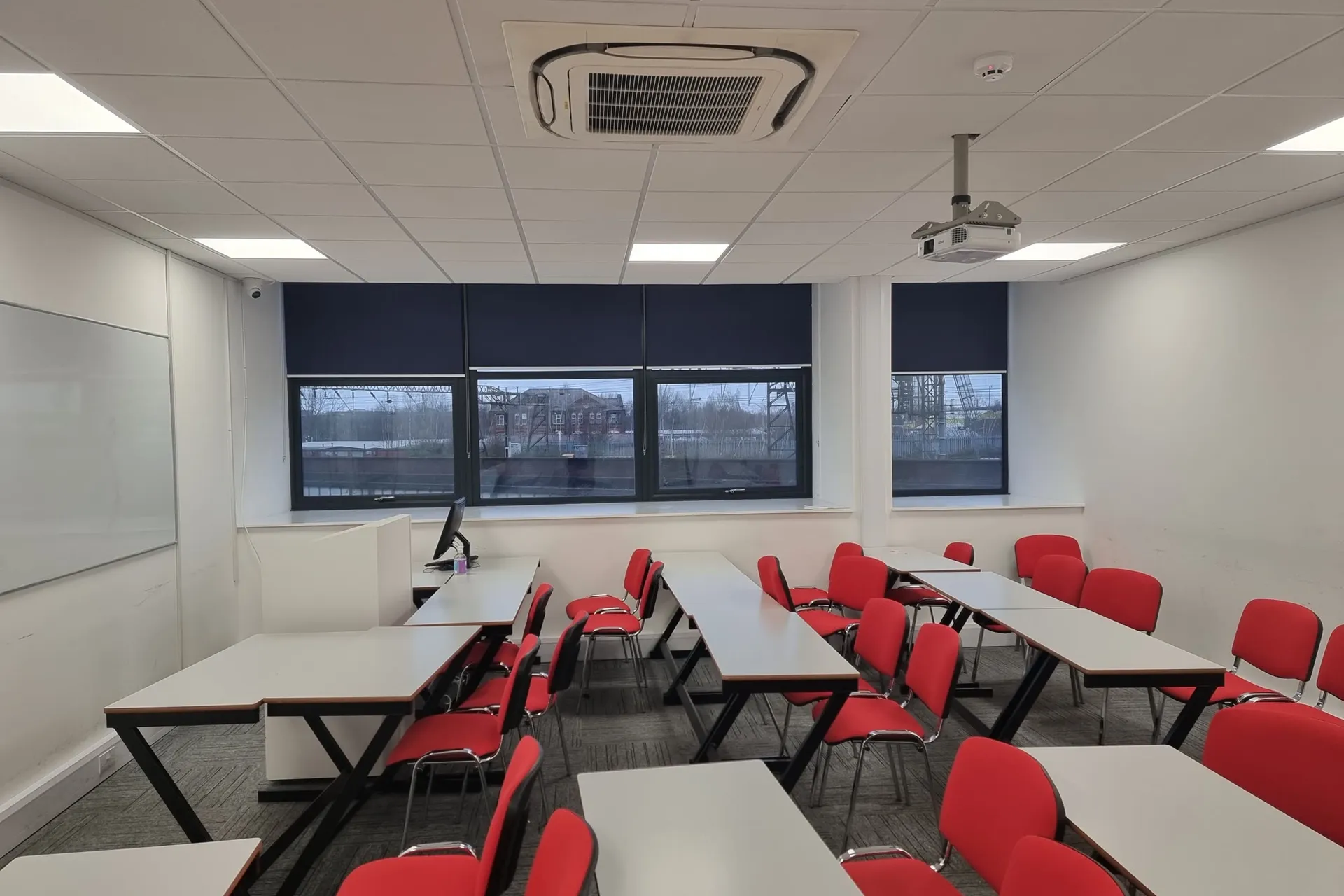 Navy roller blinds across the windows of a Global Banking School classroom in Universal Square, Manchester, with rows of red chairs and a ceiling projector