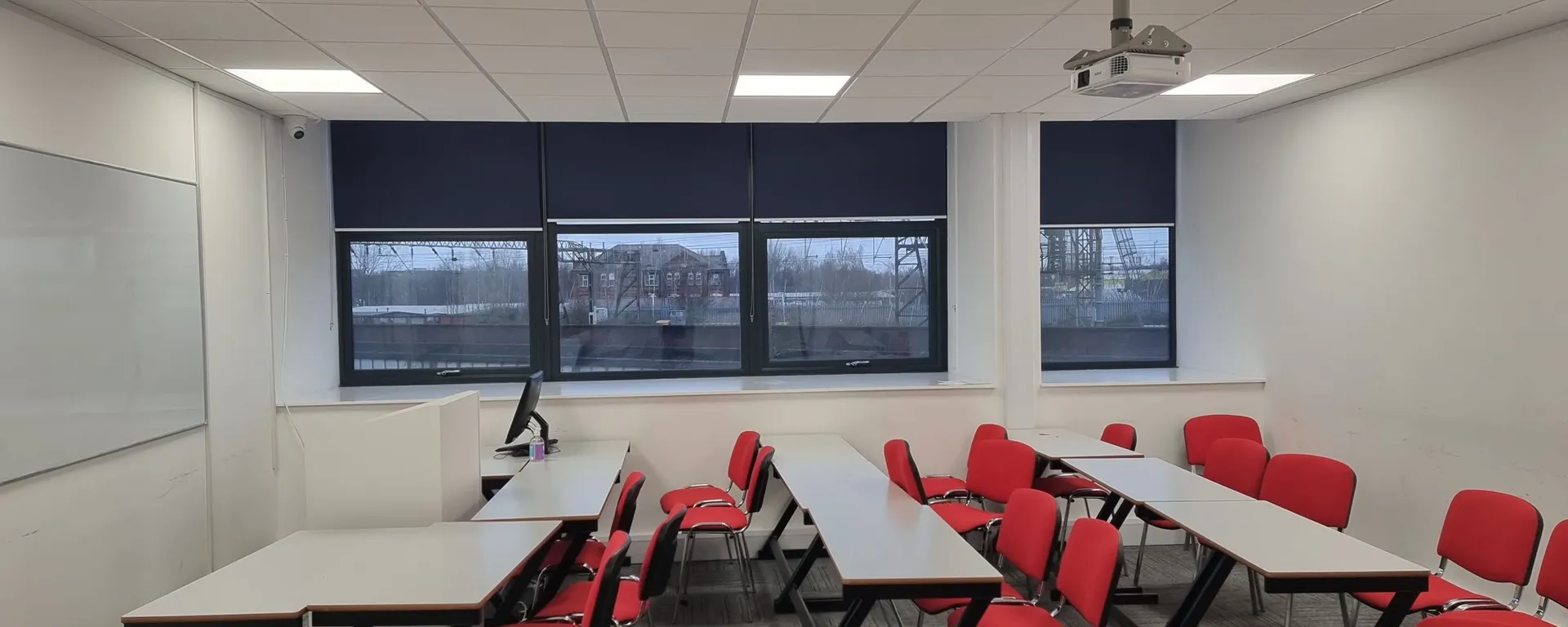Navy roller blinds across the windows of a Global Banking School classroom in Universal Square, Manchester, with rows of red chairs and a ceiling projector