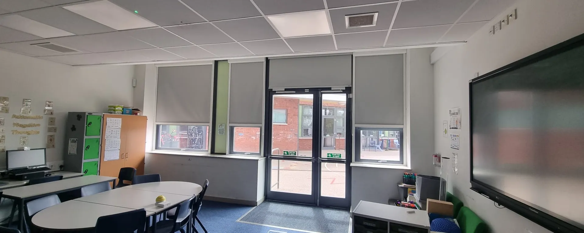 Grey roller blinds across classroom windows and the glazed external doors at The Grange Academy, oval tables in the foreground