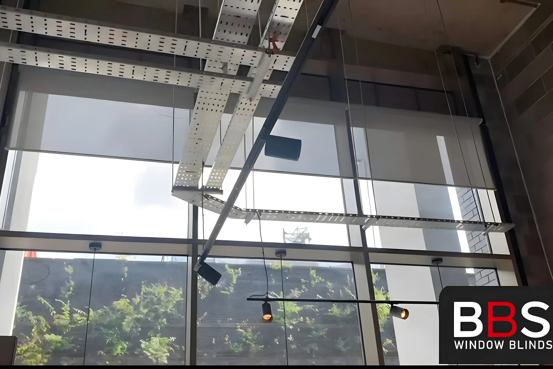 Screen fabric roller blinds lowered across the full-height glazing at Inner West cafe in Salford, cable tray and pendant lights in the ceiling above