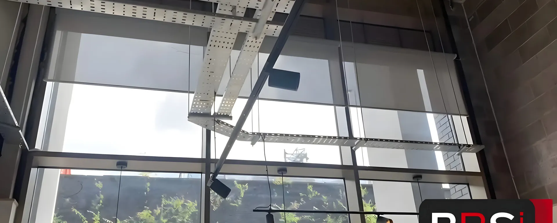 Screen fabric roller blinds lowered across the full-height glazing at Inner West cafe in Salford, cable tray and pendant lights in the ceiling above