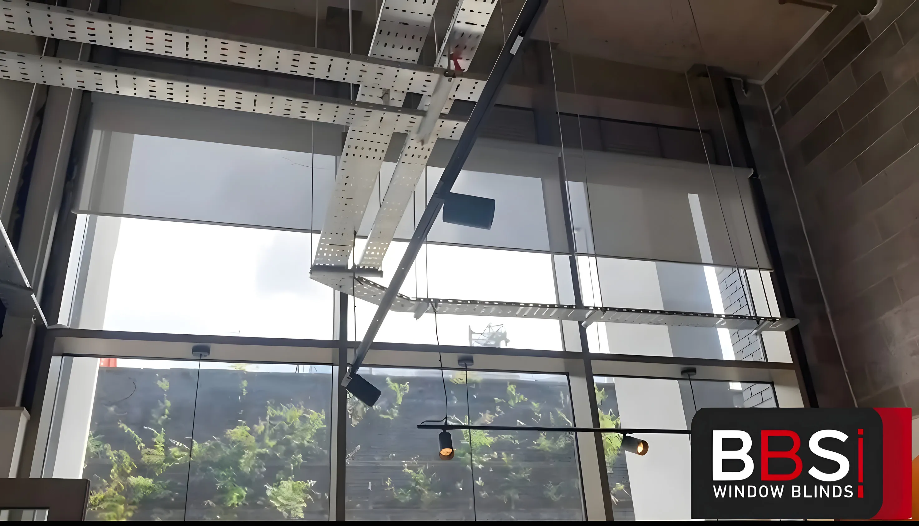 Screen fabric roller blinds lowered across the full-height glazing at Inner West cafe in Salford, cable tray and pendant lights in the ceiling above