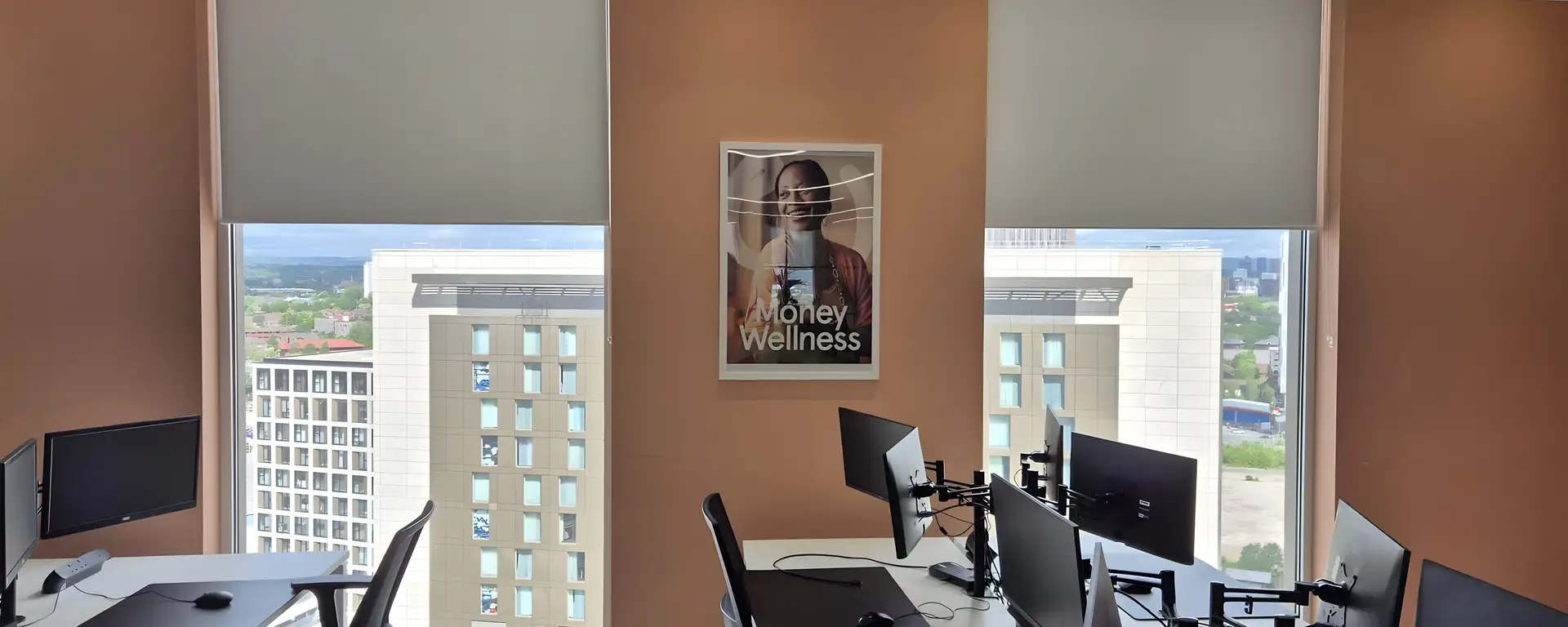 Pale dim-out roller blinds lowered over the top half of the full-height glazing at the Money Wellness office, Salford Quays, desks with monitors in front and the city beyond
