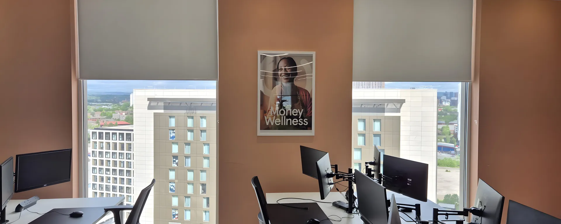 Screen fabric roller blinds half lowered across the full-height glazing of the Money Wellness office at Salford Quays, desks with monitors in front and the Quays skyline beyond