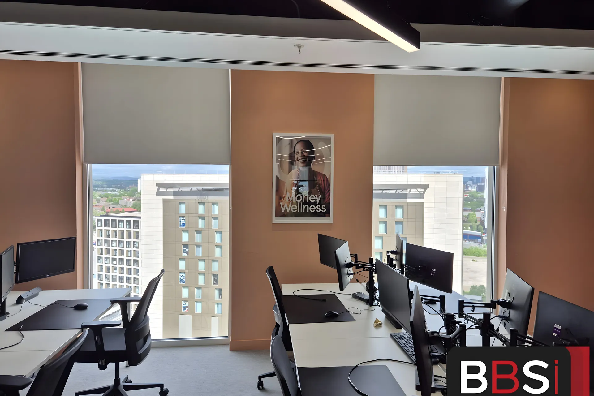 Screen fabric roller blinds half lowered across the full-height glazing of the Money Wellness office at Salford Quays, desks with monitors in front and the Quays skyline beyond
