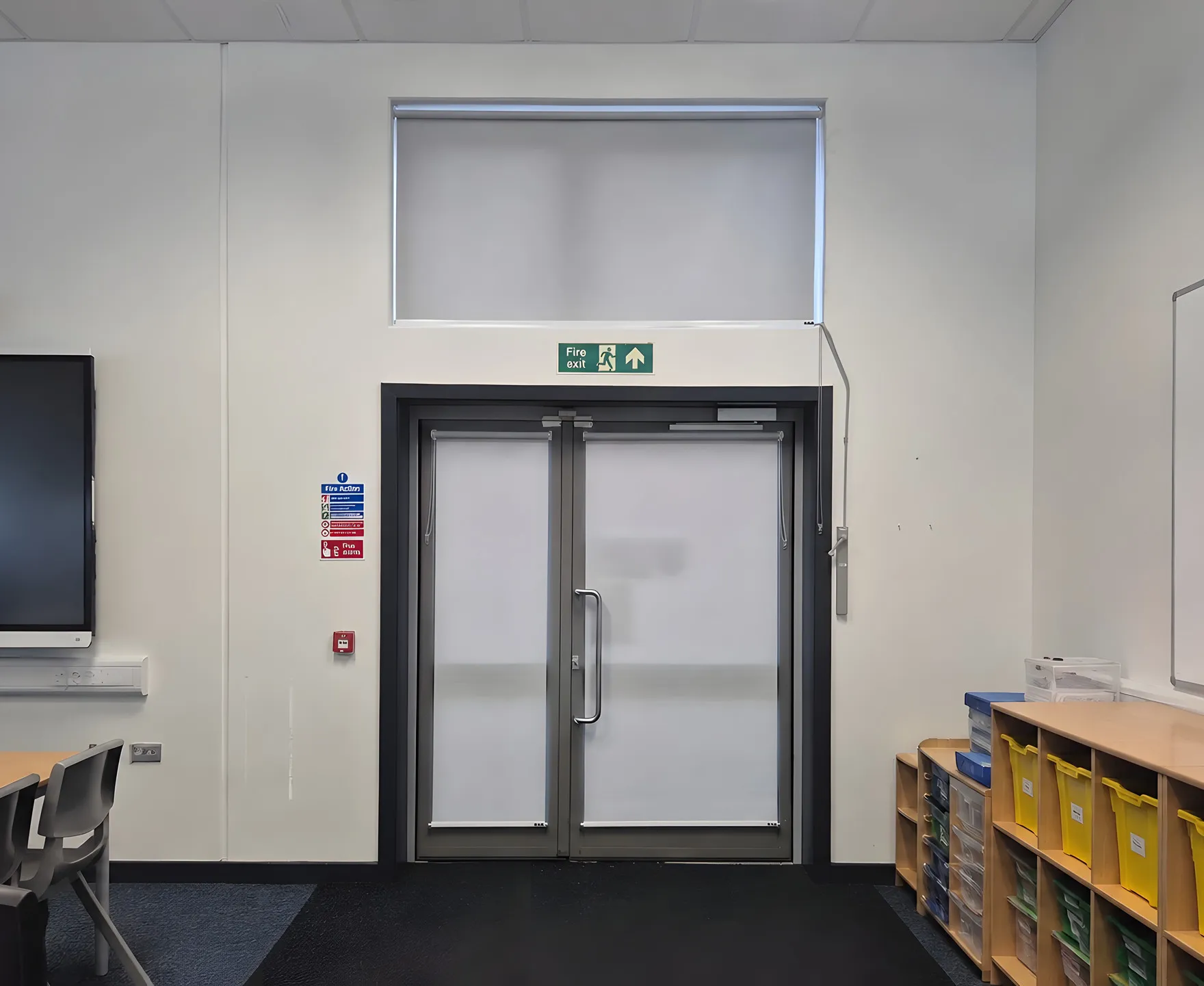 Roller blinds on both leaves of a fire exit door and on the overhead panel, fitted around the door closers