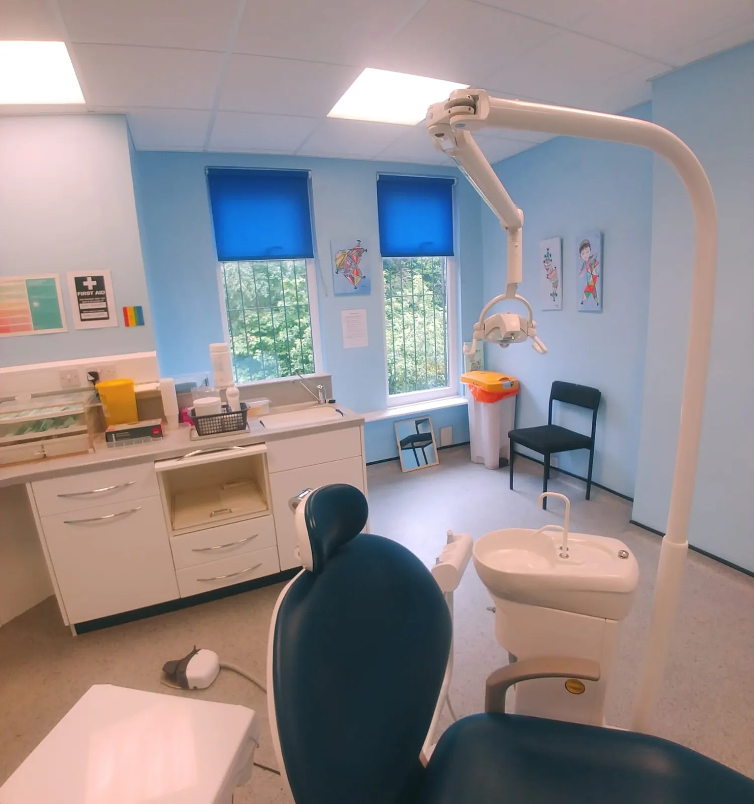 Two bright blue roller blinds in a dental treatment room, with children's artwork on the wall between the windows