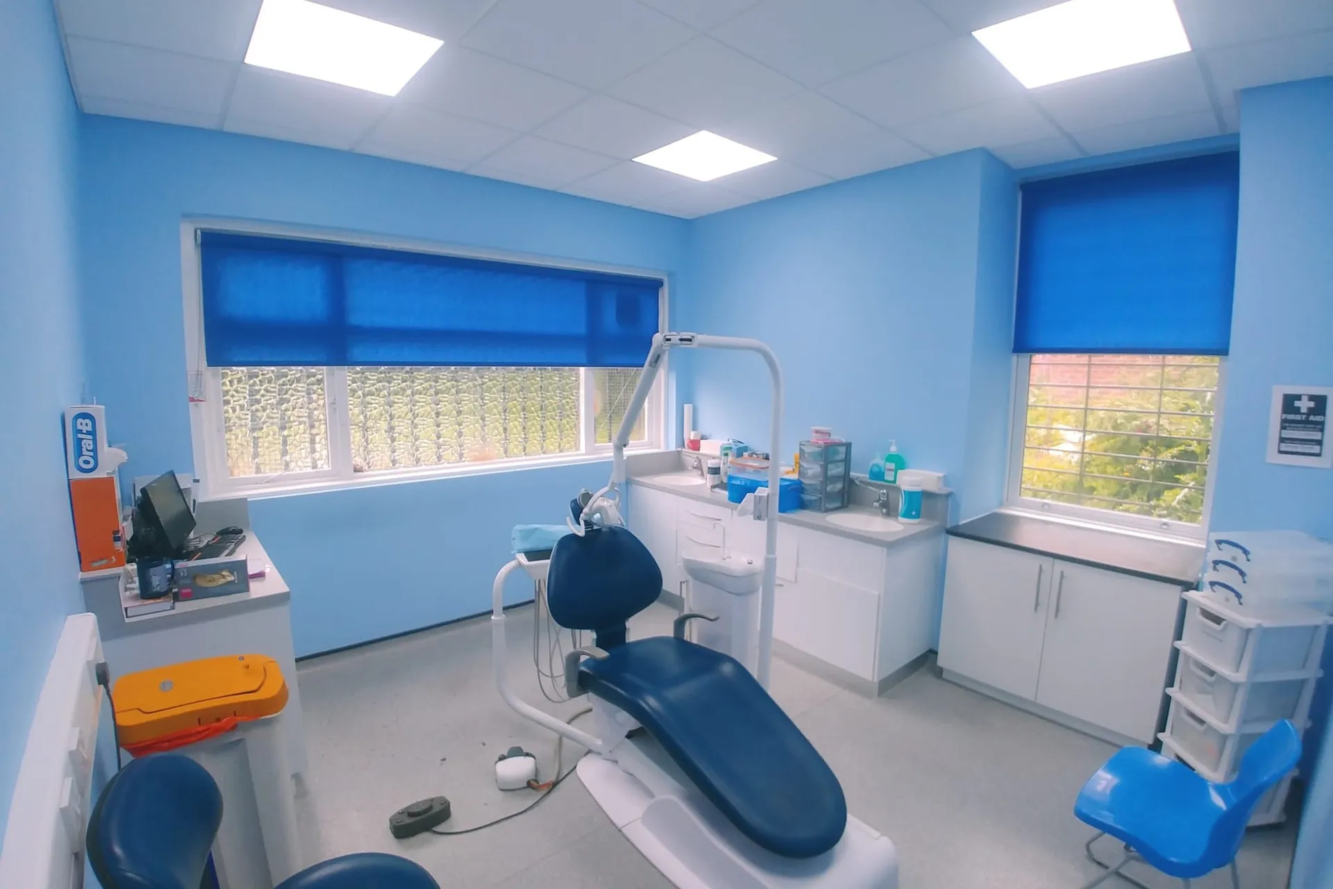 Bright blue roller blinds on two treatment-room windows at a dental surgery in North Manchester, with the dental chair in the centre