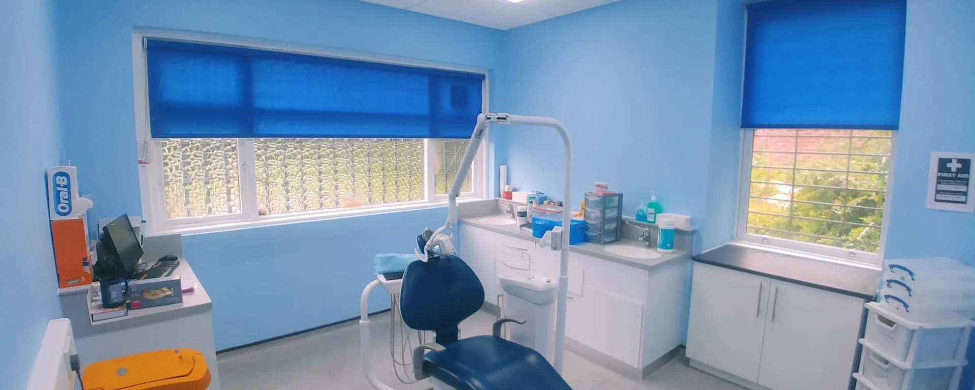 Bright blue roller blinds on two treatment-room windows at a dental surgery in North Manchester, with the dental chair in the centre