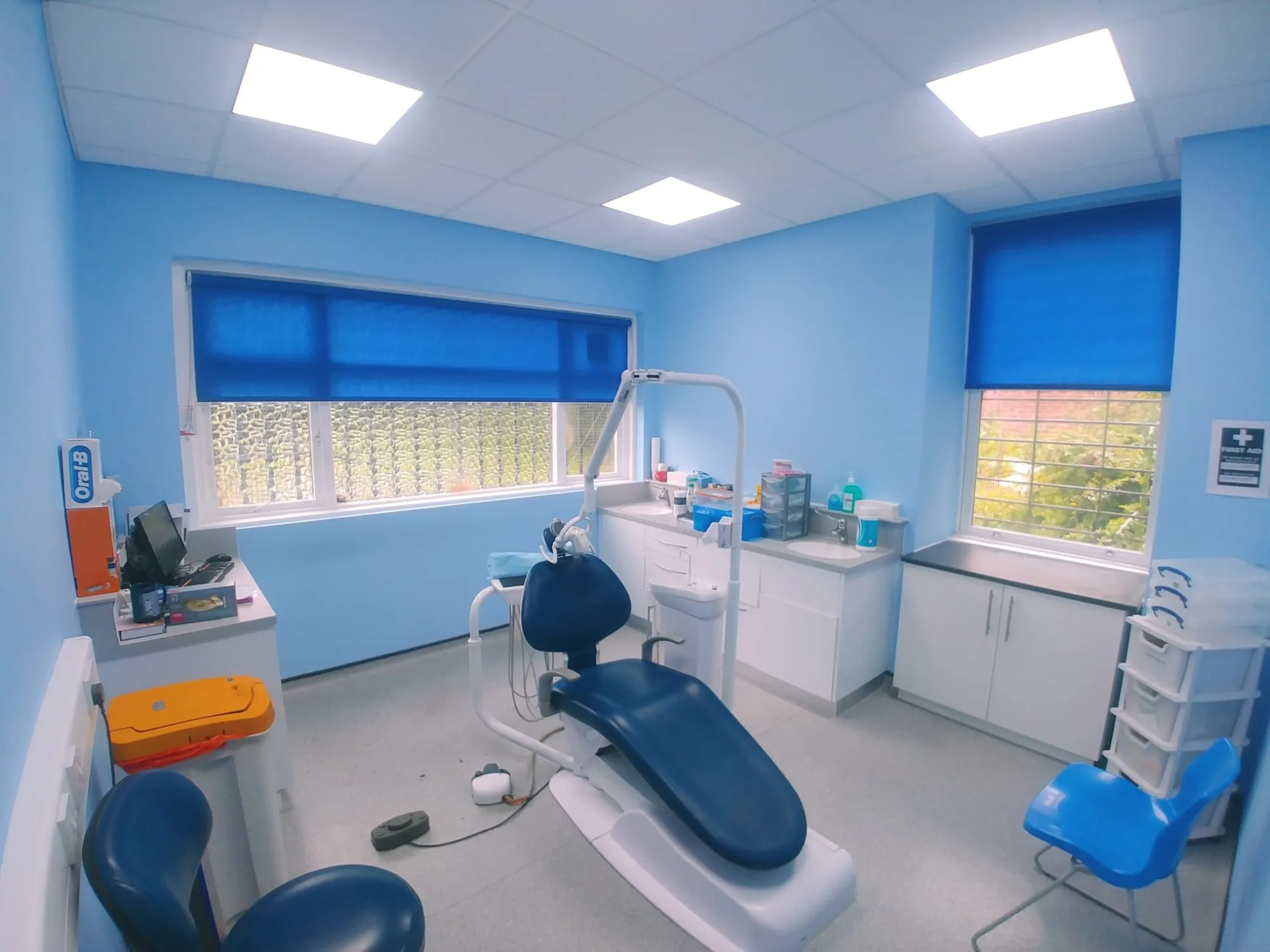 Bright blue roller blinds on two treatment-room windows at a dental surgery in North Manchester, with the dental chair in the centre