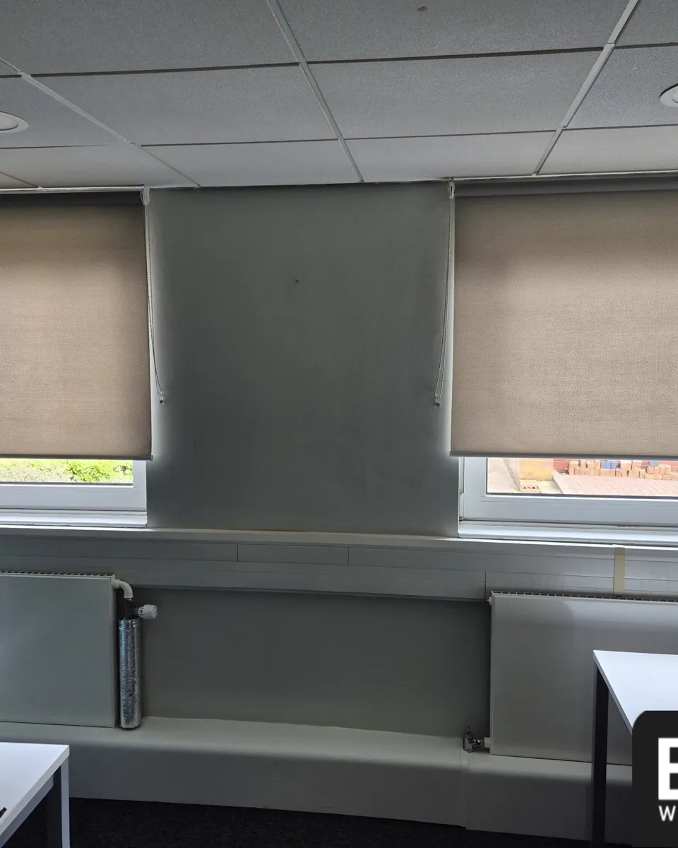 Two neutral fabric roller blinds half lowered over office windows at North West Ambulance Service headquarters, radiators beneath