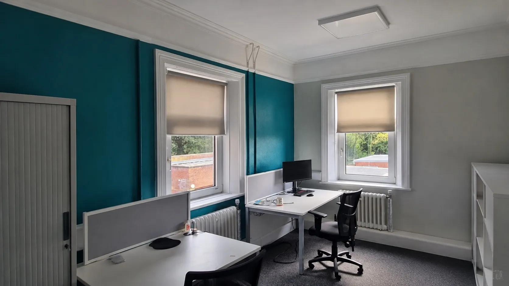 Corner office at NWAS HQ with roller blinds part lowered over the two tall windows above the desks