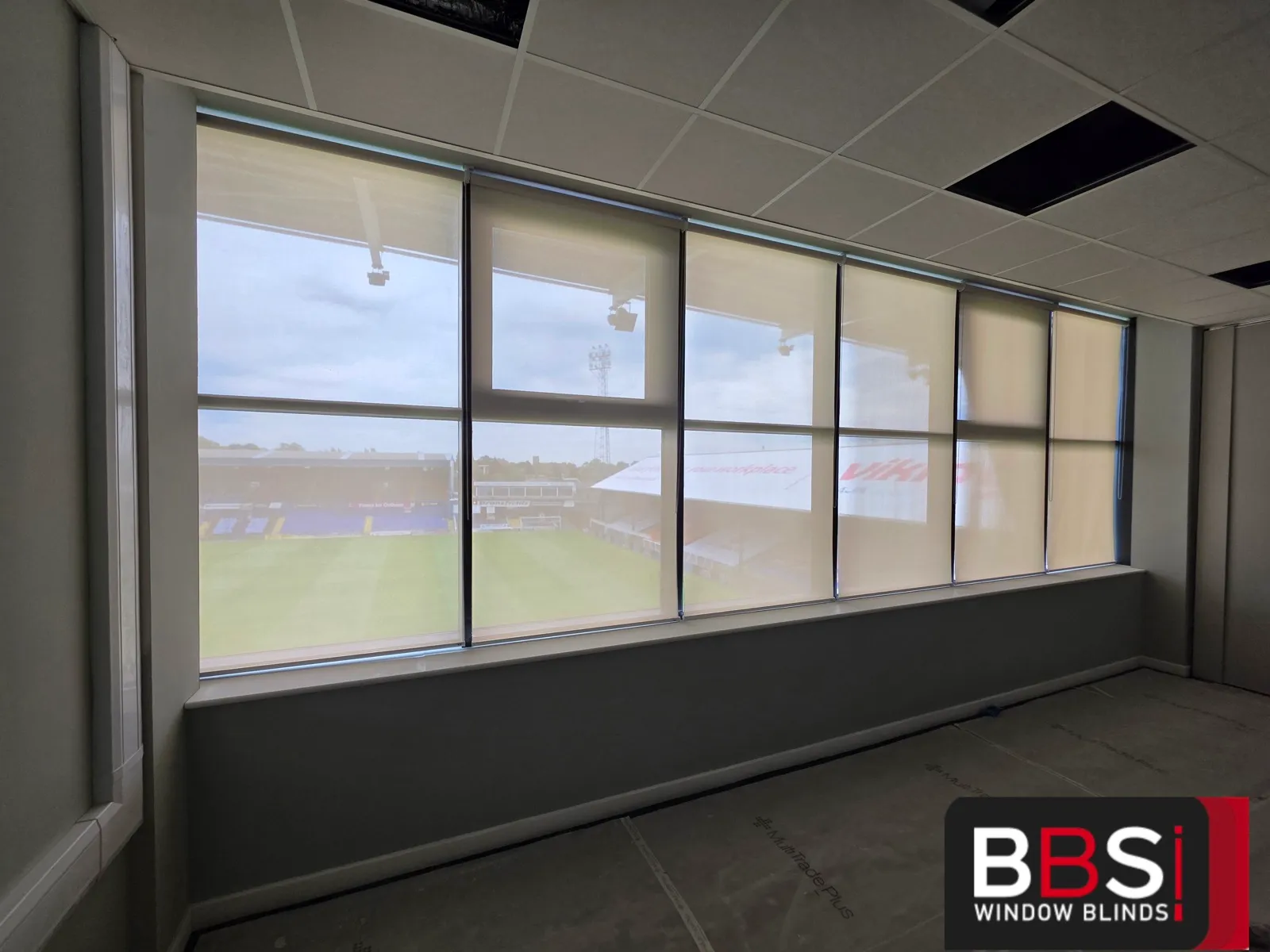 Screen fabric blinds fully lowered across a refurbished room at Boundary Park, daylight softened and the pitch still readable through the fabric