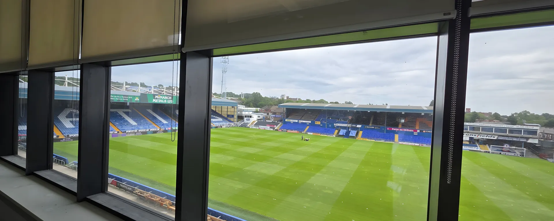 Screen fabric roller blinds part lowered across a bank of windows at Boundary Park, the pitch and opposite stand in full view below