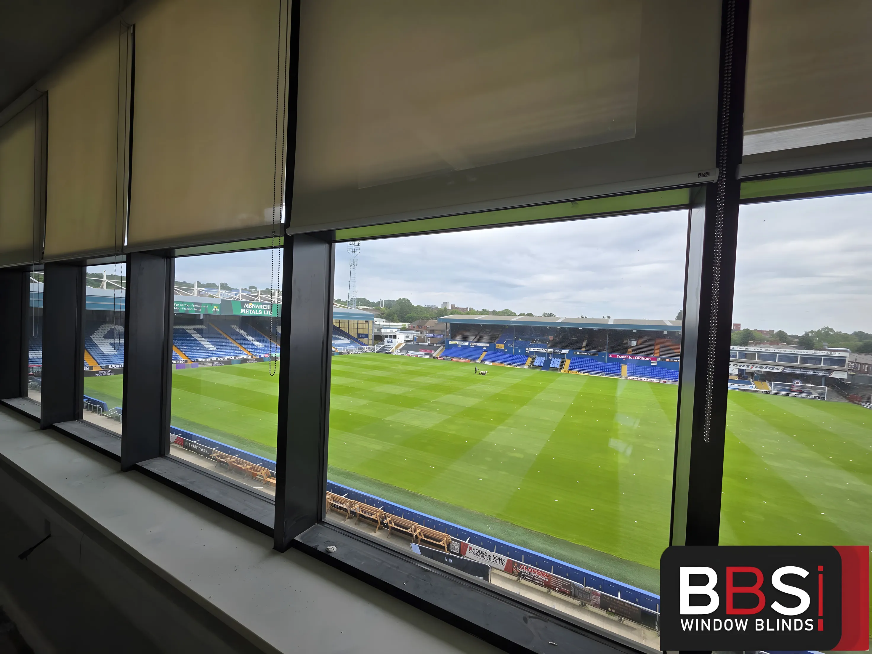 Screen fabric roller blinds part lowered across a bank of windows at Boundary Park, the pitch and opposite stand in full view below