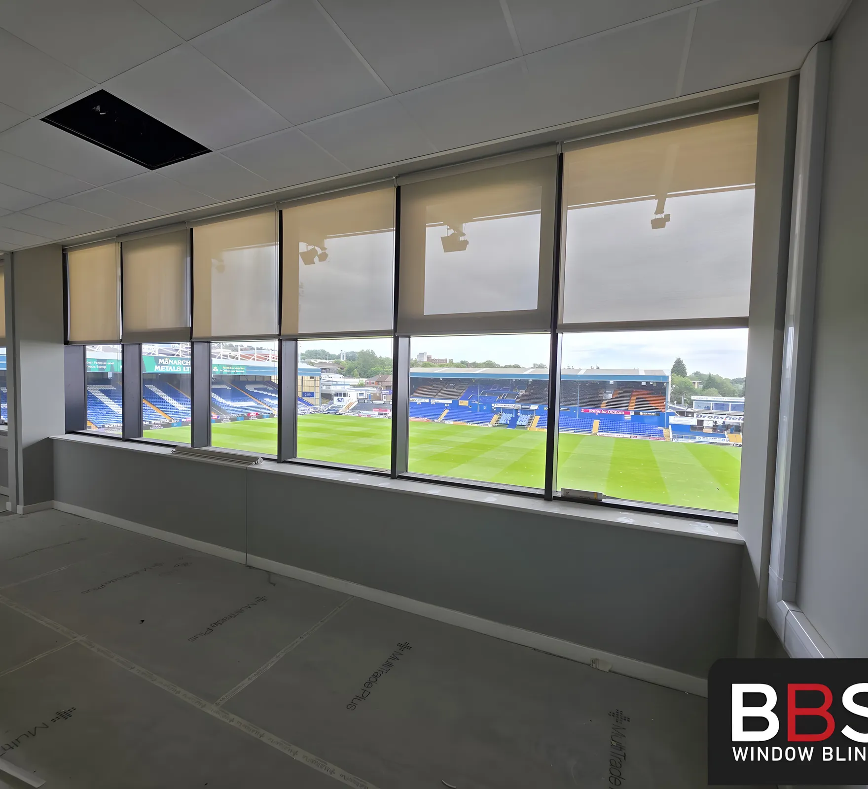 A long run of screen fabric blinds down the pitch-facing elevation at Boundary Park, pitch visible through the glazing