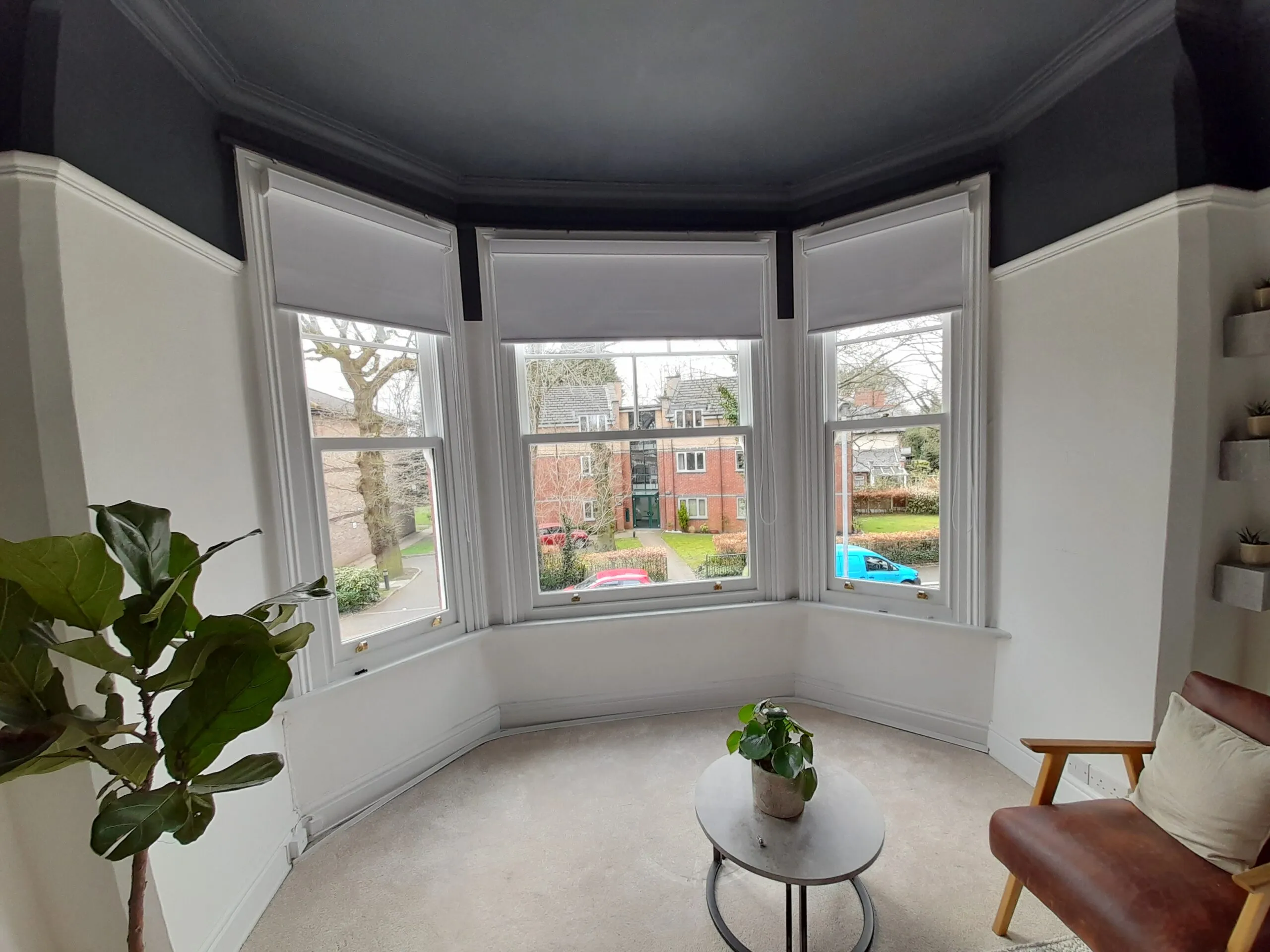 Grey roller blinds fitted across a bay window in a Manchester living room, armchair and plants beside it