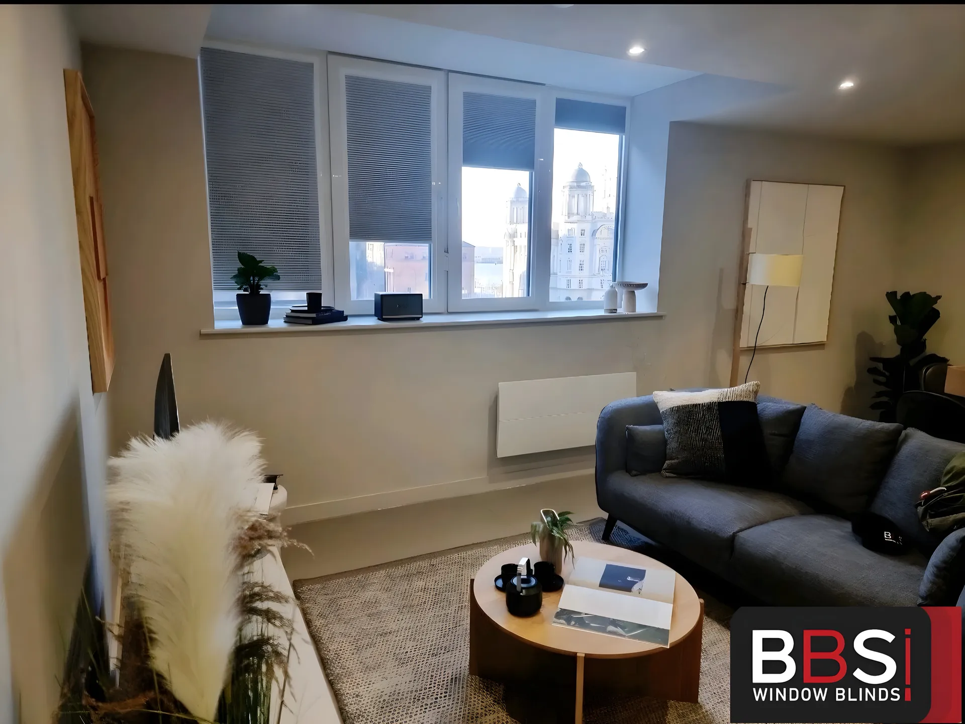Grey perfect fit pleated blinds across a three-pane window in a Rocco House apartment lounge, with Liverpool waterfront buildings beyond the glass