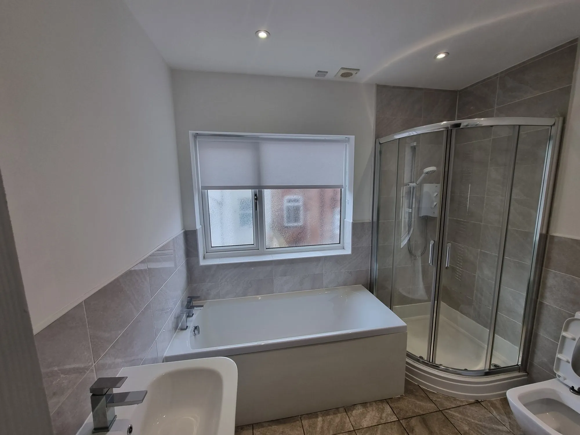 Grey roller blind at a bathroom window between the bath and a walk-in shower