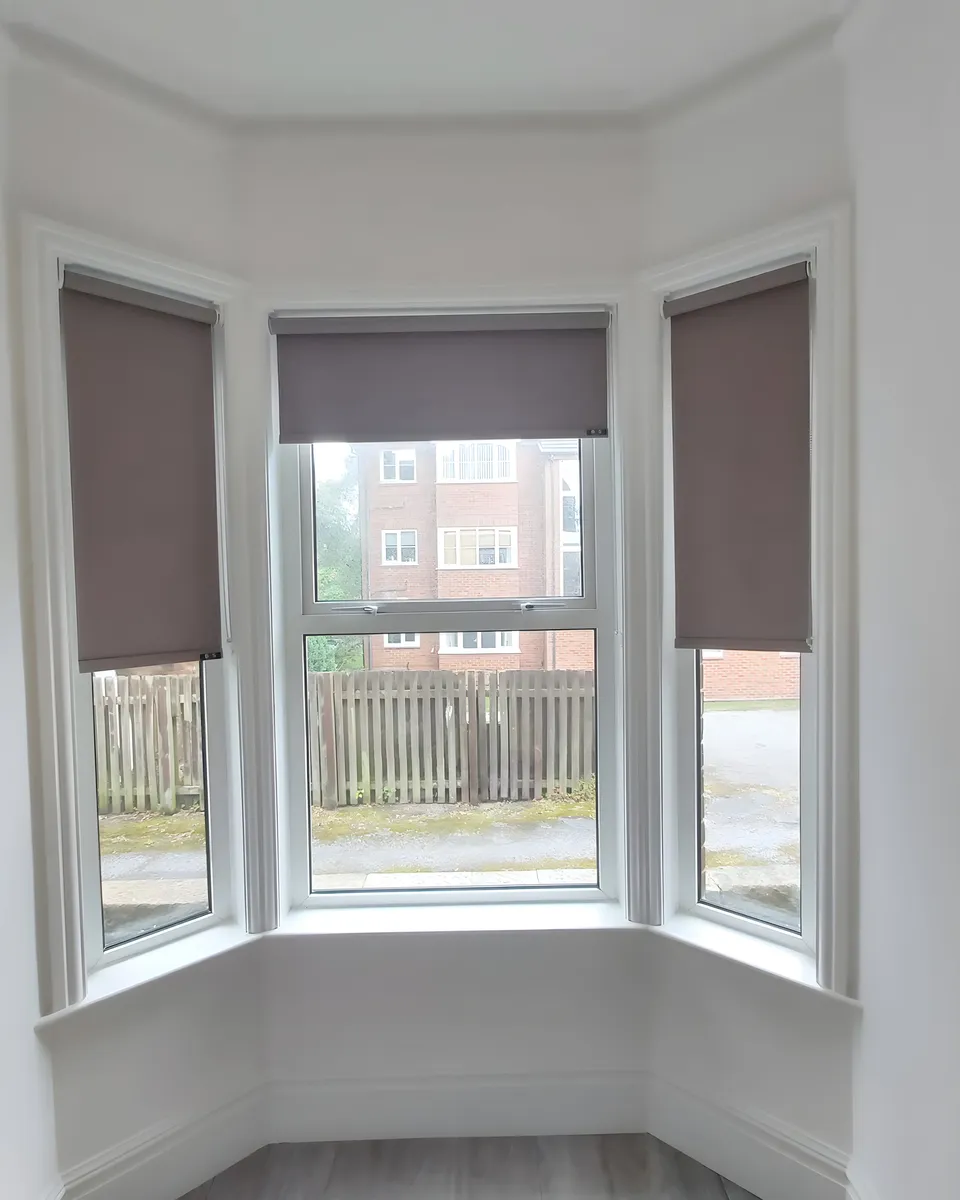 Taupe roller blinds fitted inside each of the three panes of a white bay window with cornicing above