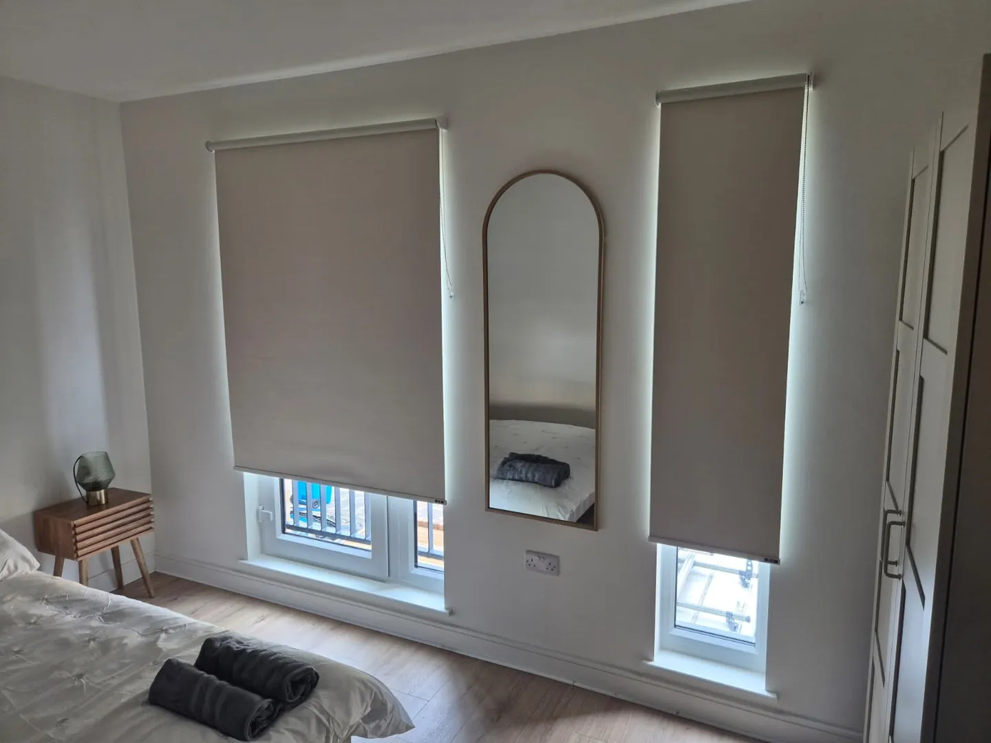 Cream roller blinds on a wide and a narrow bedroom window with a gold arched full-length mirror between them