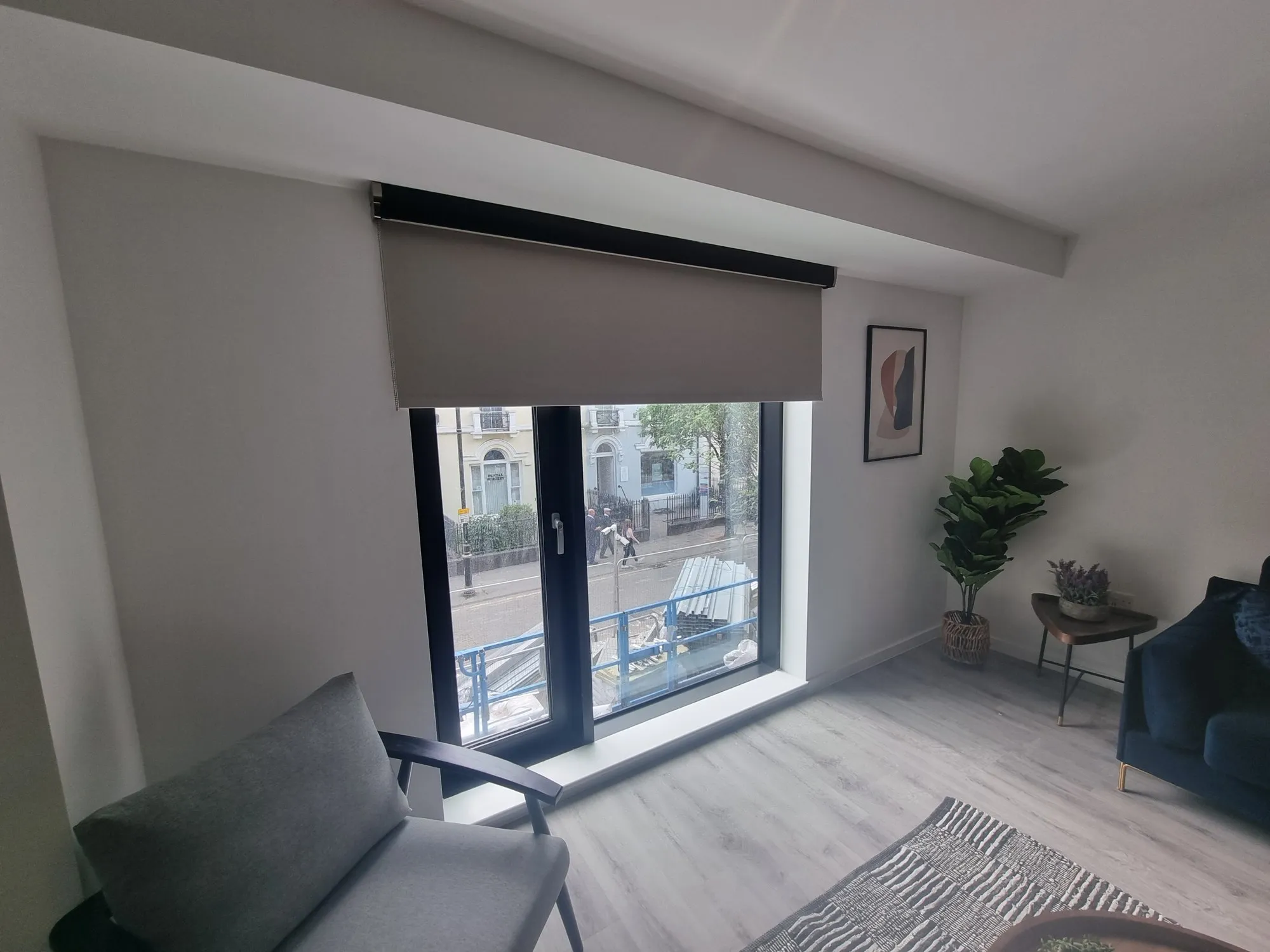 Grey roller blind lowered over balcony doors in a bedroom with a plant beside the bed