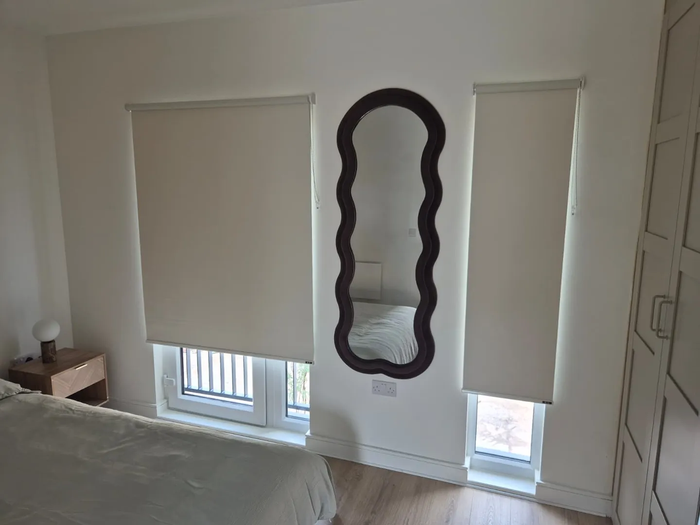 Cream roller blinds on a wide and a narrow bedroom window with a dark wavy full-length mirror between them